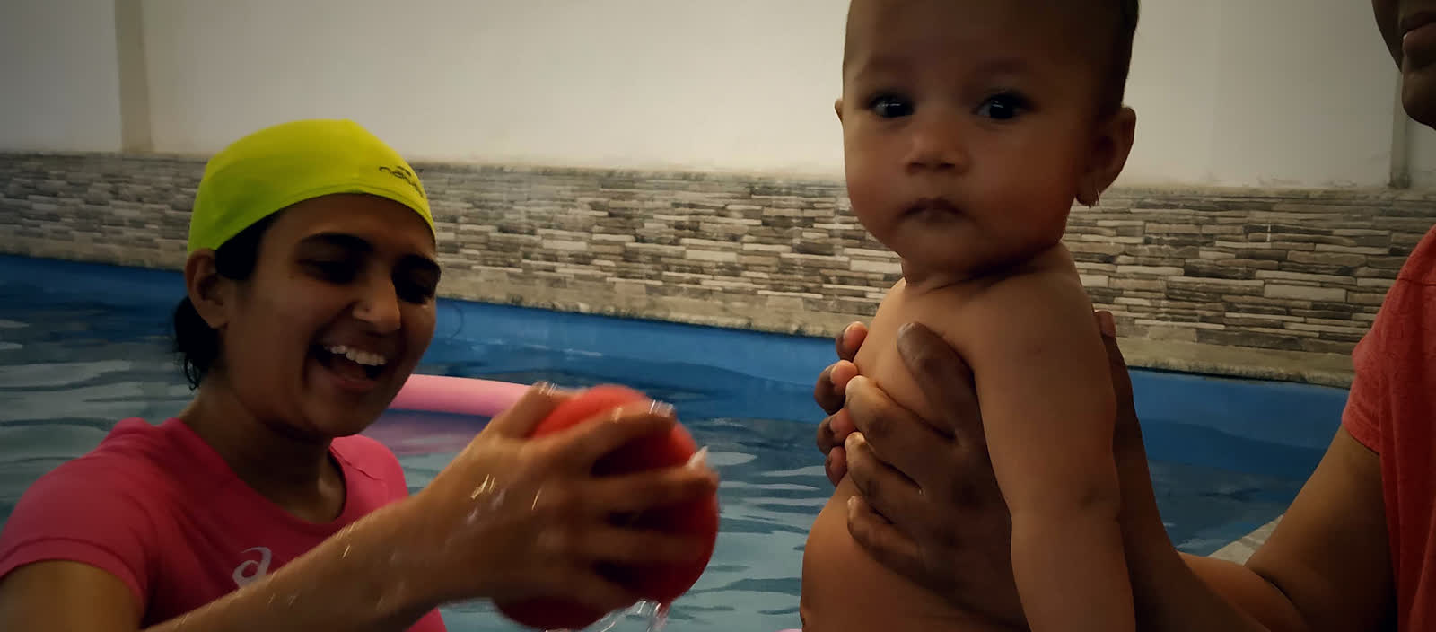 A therapist plays with a baby at the edge of the warm pool, pouring water from a soft ball while a parent supports the child